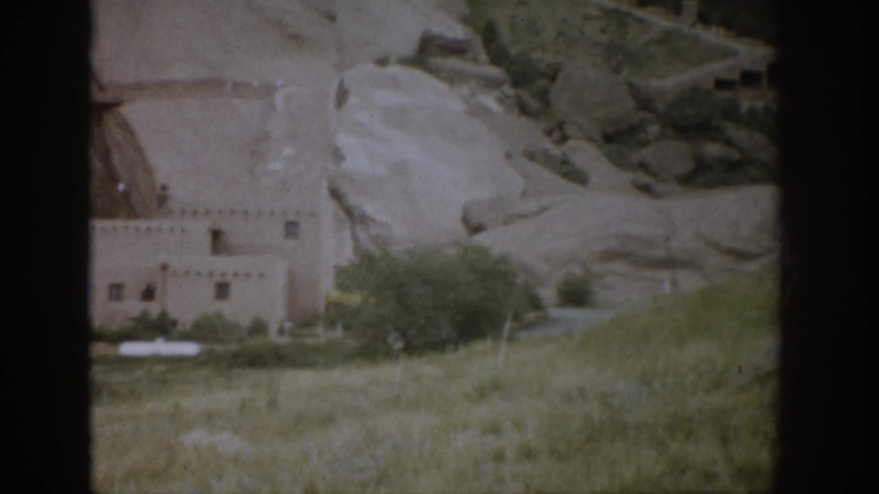 Adobe-style building below pale rock cliffs with grass and shrubs in front.