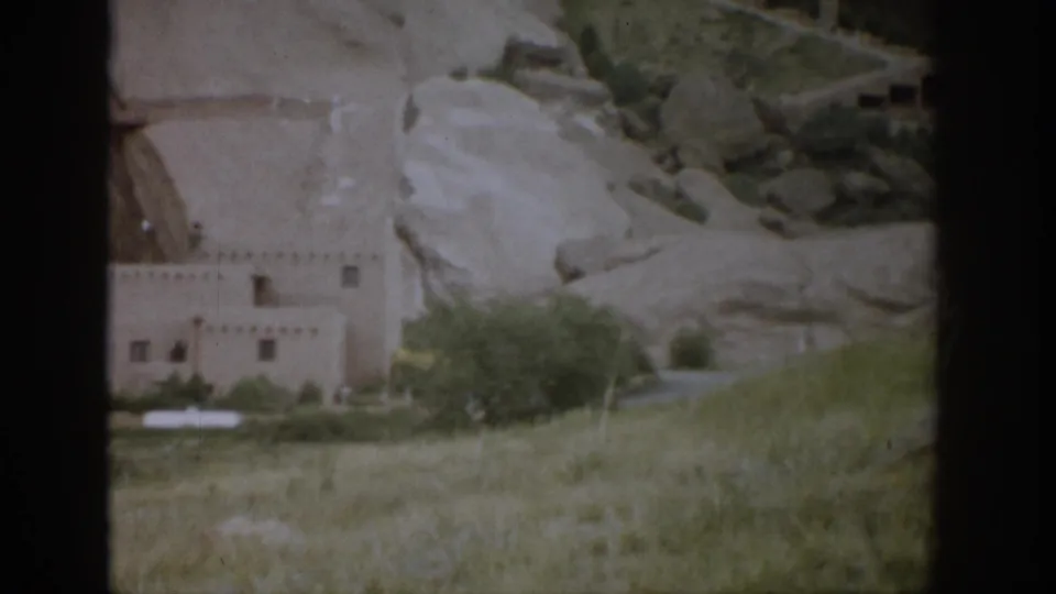 1959 Colorado 8mm frame showing an adobe-style building at the base of pale rocky cliffs with open grass and shrubs.