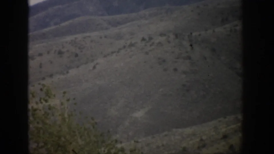 1959 Colorado home-movie frame showing open dry mountain terrain with scattered shrubs and distant ridges.