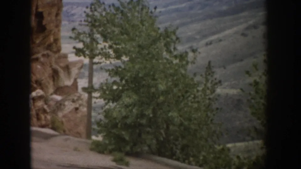 1959 Colorado home-movie still with a rugged rock edge, foreground tree, and hillside vista beyond.