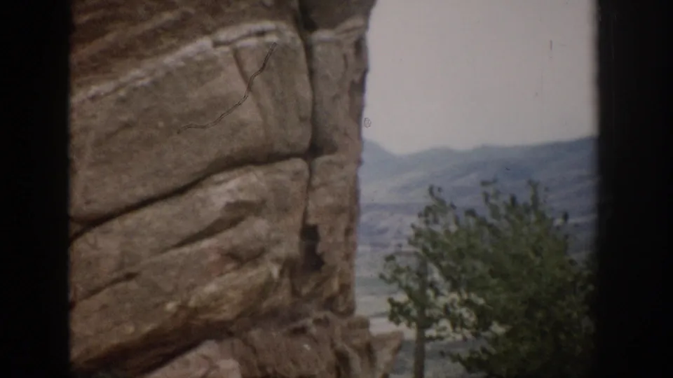 1959 Colorado 8mm frame showing tall layered rock formations with a tree and distant mountains visible through a gap.