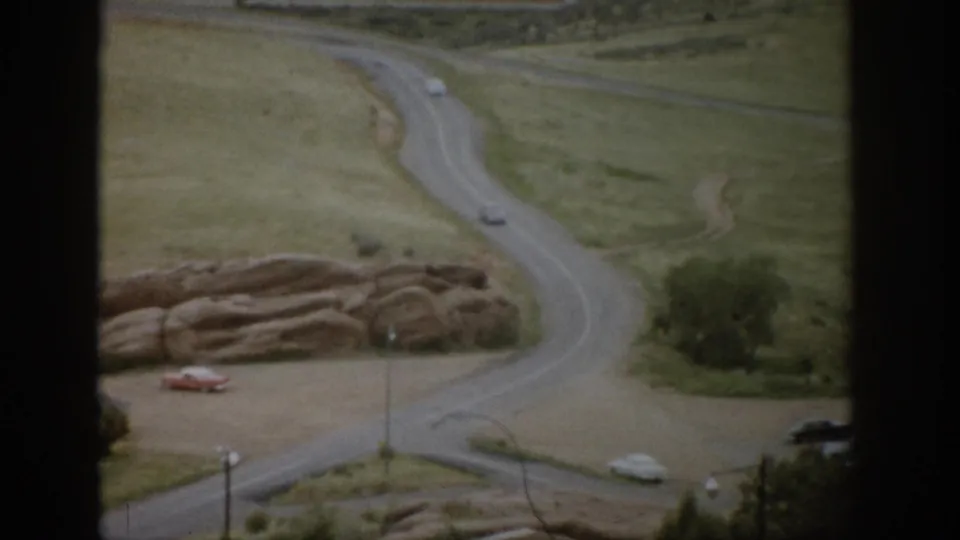 1959 Colorado overlook view with a curving road, grassy slopes, exposed rocks, and mid-century cars below.