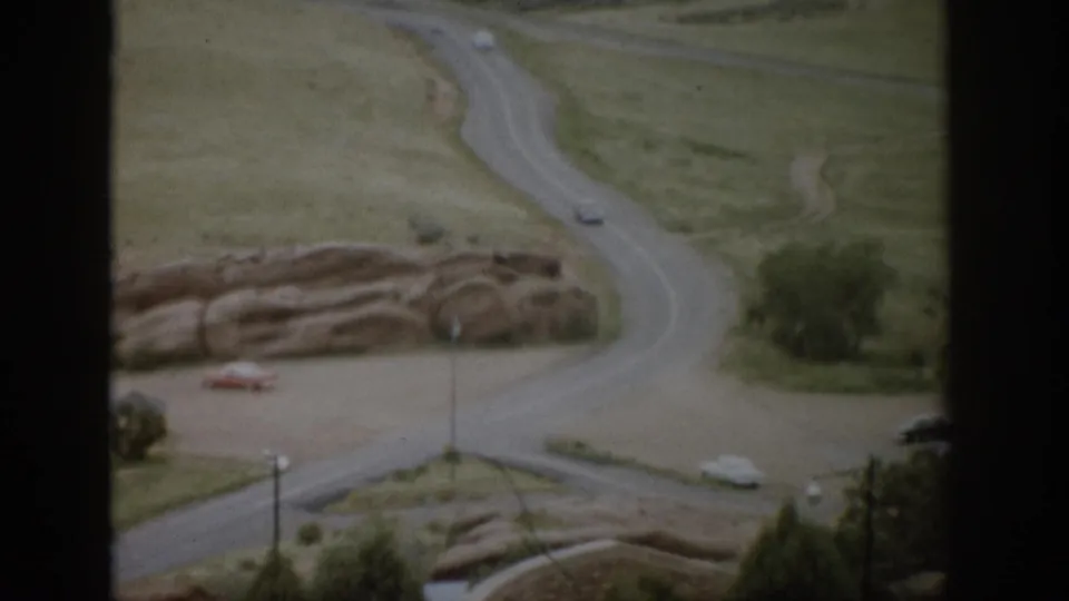 Colorado 1959 home-movie view from an overlook, showing a winding road, rock outcrops, grass, shrubs, and small cars.