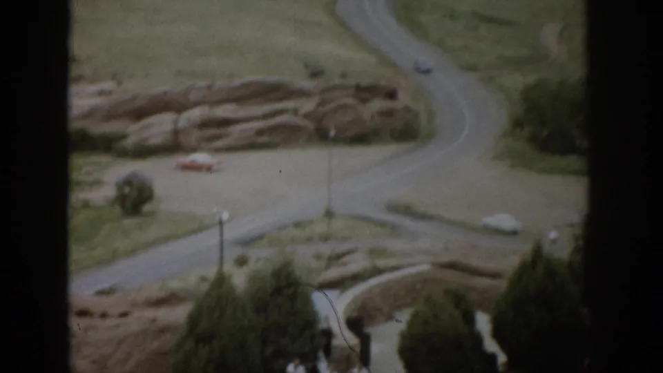 1959 Colorado home-movie view of a winding park road, rocky grassland, trees, and small cars seen through a dark frame.