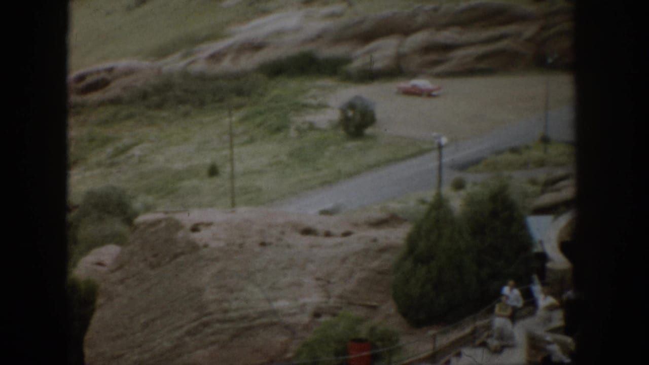 Rocky Colorado park terrain with a road, parked car, lampposts, trees, and distant tourists partly blocked by dark framing.