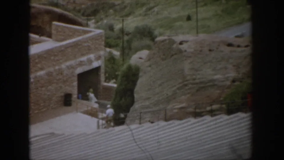 1959 Colorado home-movie frame showing a stone building, amphitheater seating, paths, trees, rocky slopes, and distant tourists.