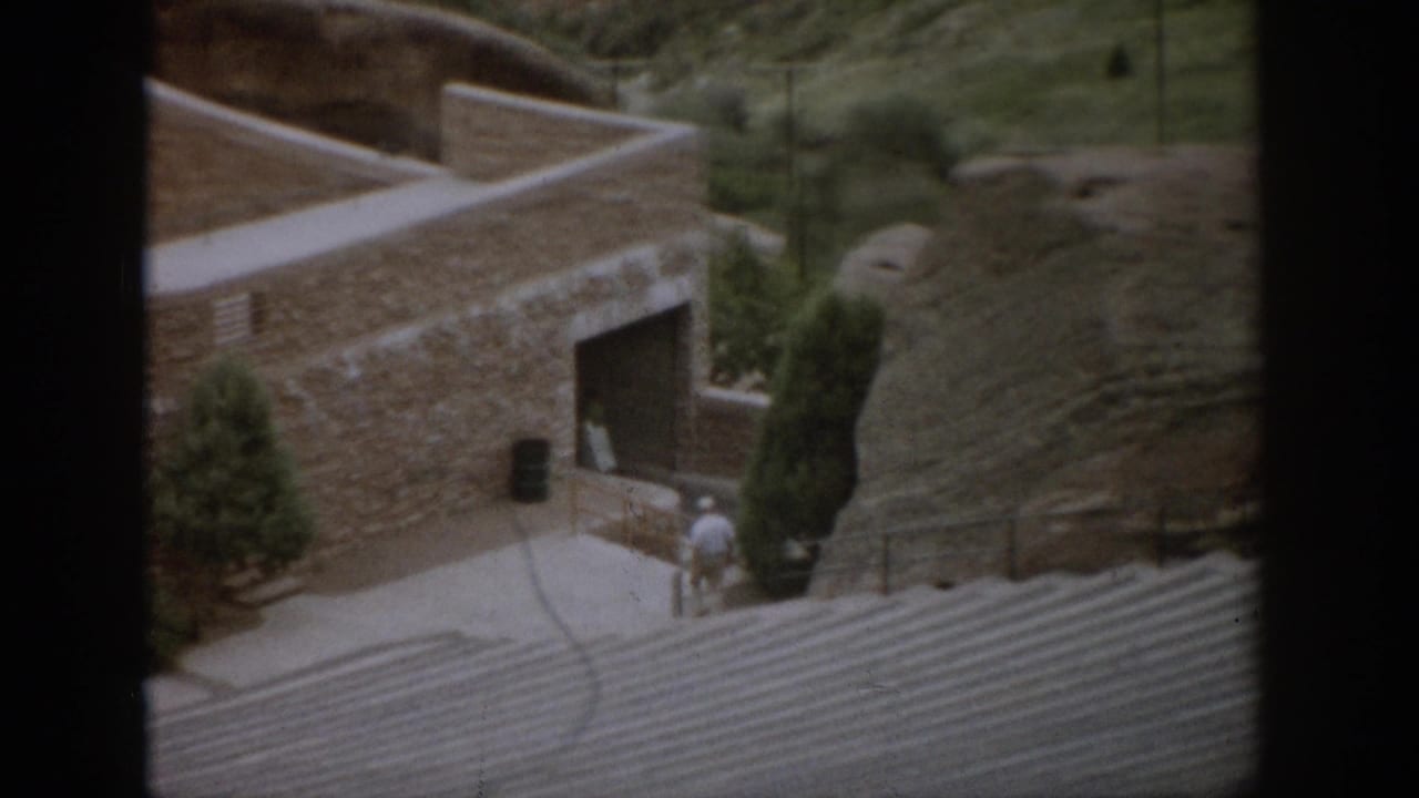 1959 Colorado 8mm frame showing a stone hillside building with curved walls, walkways, steps, small evergreens, and visitors by the entrance.