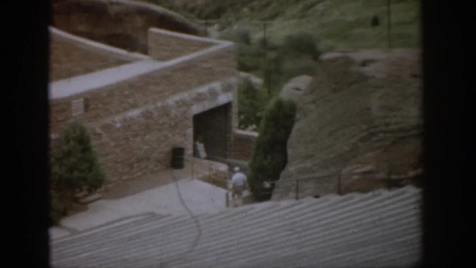 1959 Colorado 8mm frame showing a stone hillside building with curved walls, walkways, steps, small evergreens, and visitors by the entrance.