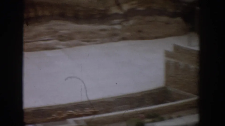 Soft 1959 Colorado 8mm frame showing a pale concrete path, stone wall, rocky hillside, and visible film marks during the pan.