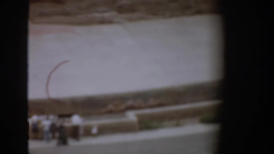 1959 Colorado 8mm frame with visitors near a low wall, a pale walkway, curved stonework, trees, and hillside terrain.