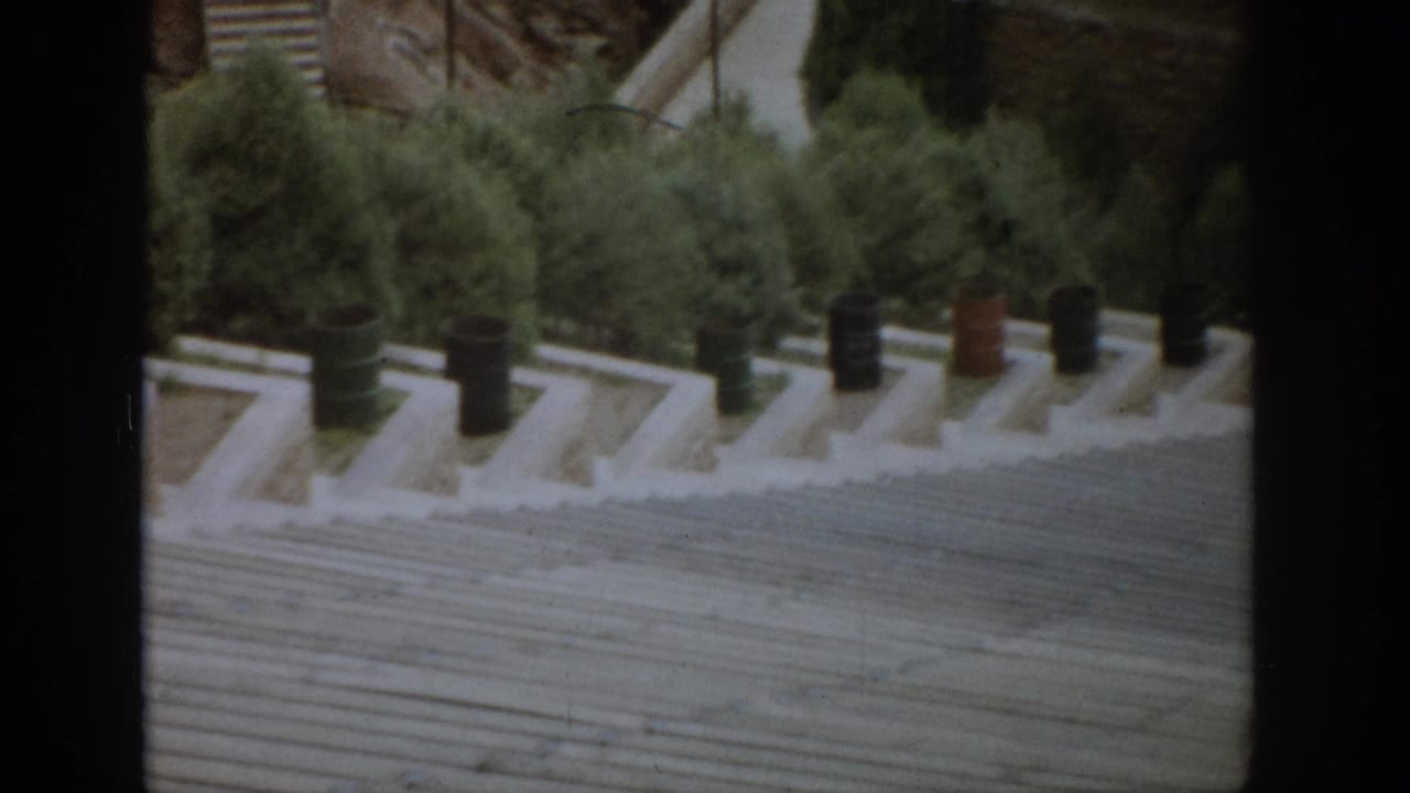 1959 Colorado home-movie frame with an angled view across a broad stone stairway, barrel decorations, and dense evergreens.