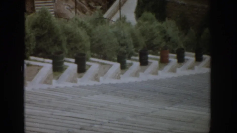 1959 Colorado home-movie frame with an angled view across a broad stone stairway, barrel decorations, and dense evergreens.