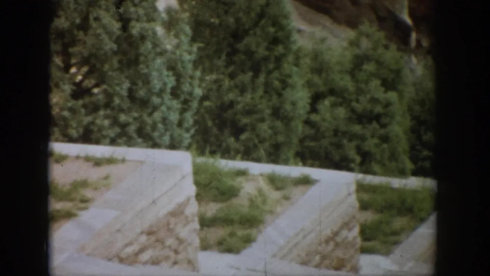 1959 Colorado 8mm frame showing a stone retaining wall, pale edging, grass, and dense green trees from a visitor viewpoint.
