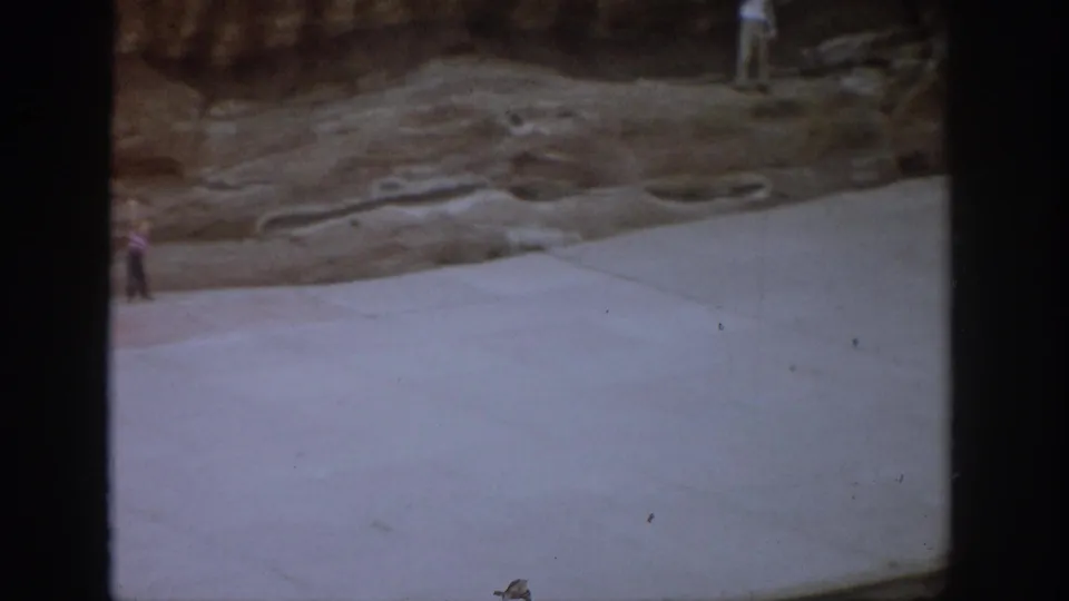Colorado 1959 home-movie frame showing a rock-lined site, pale ground surface, and partially visible distant people near the frame edges.