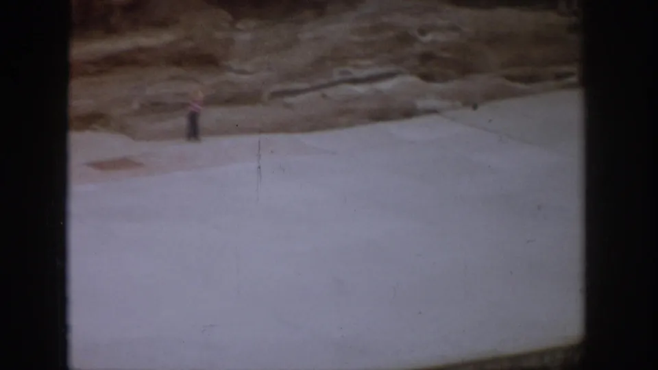 1959 Colorado 8mm frame with a small distant person on a pale platform beneath a layered rock face at an archaeological site.