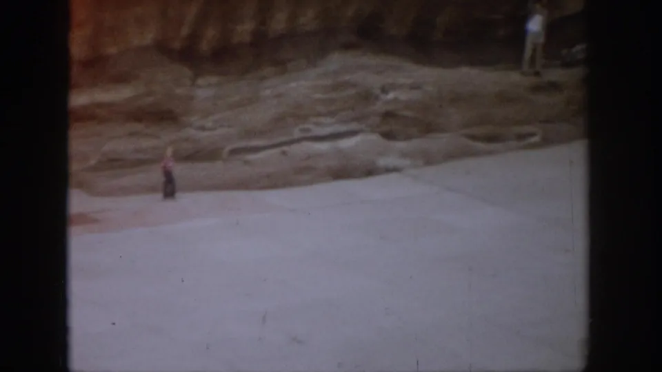1959 Colorado 8mm frame showing an open amphitheater floor, a distant visitor, and a reddish rocky backdrop.
