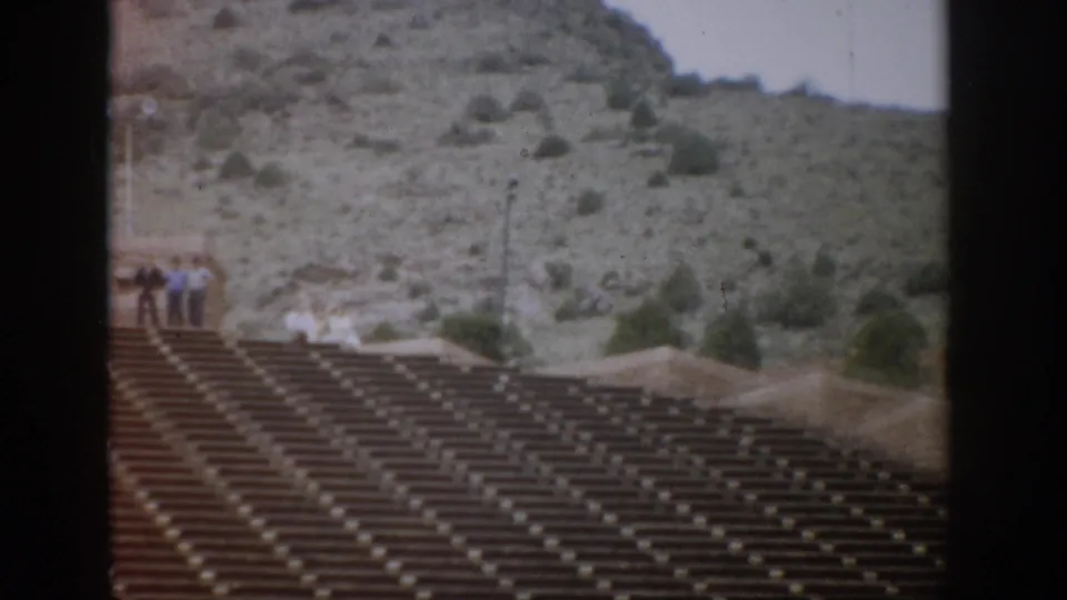 1959 Colorado frame with empty amphitheater rows, a few tourists at the upper edge, and scrub-covered rock slopes behind them.