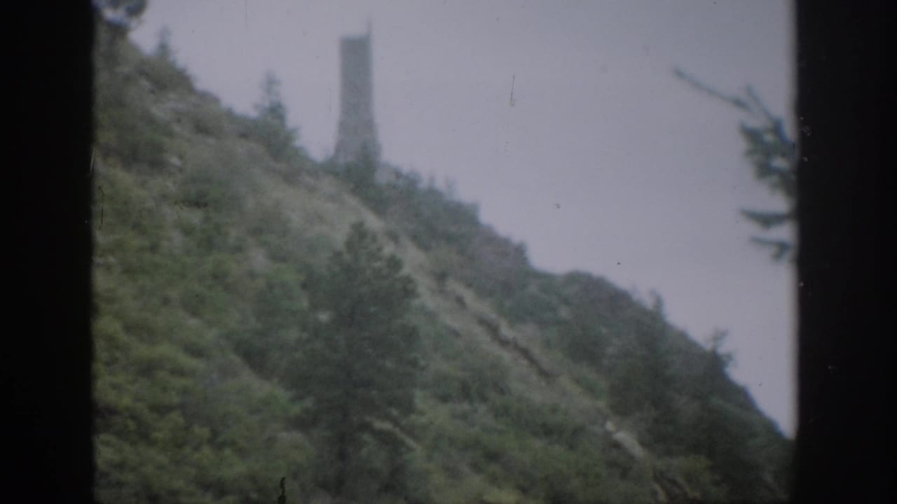 1959 Colorado home-movie frame showing a steep green hillside, gray sky, and a tall distant tower-like structure seen through dark window framing.