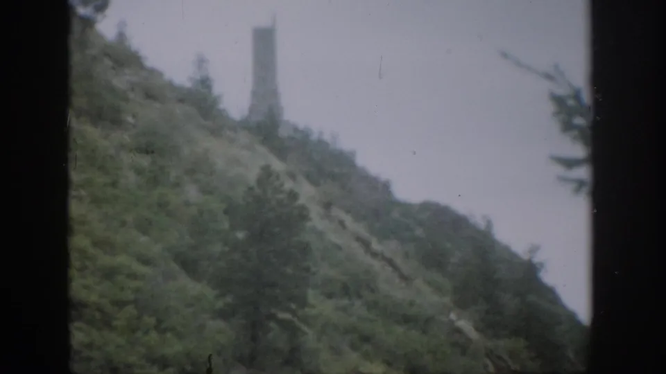 1959 Colorado home-movie frame showing a steep green hillside, gray sky, and a tall distant tower-like structure seen through dark window framing.