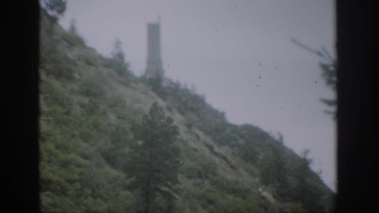 1959 Colorado 8mm frame of a brushy hillside and trees, with a pale distant tower-like structure partly softened by haze or focus.