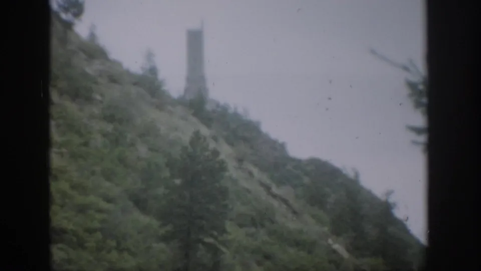1959 Colorado 8mm frame of a brushy hillside and trees, with a pale distant tower-like structure partly softened by haze or focus.