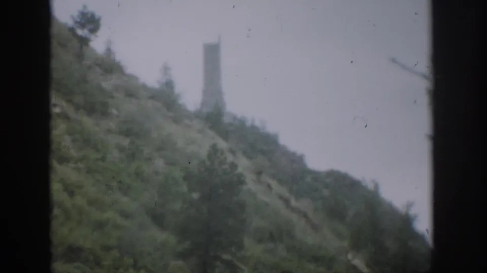 Soft 1959 Colorado home-movie view of a steep wooded hillside, with a distant vertical tower-like shape above the ridge line.