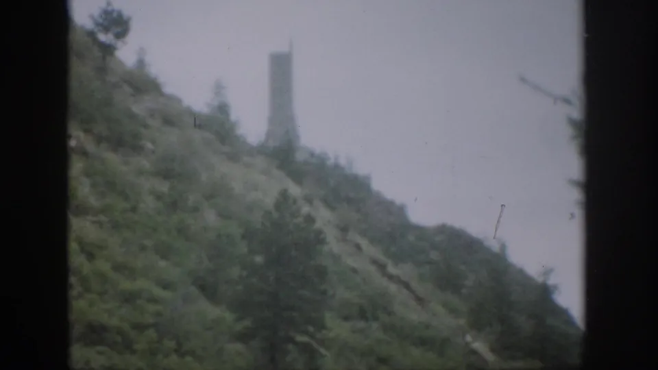 1959 Colorado frame showing the wooded slope and distant tower-like structure, with dark framing and soft home-movie grain.