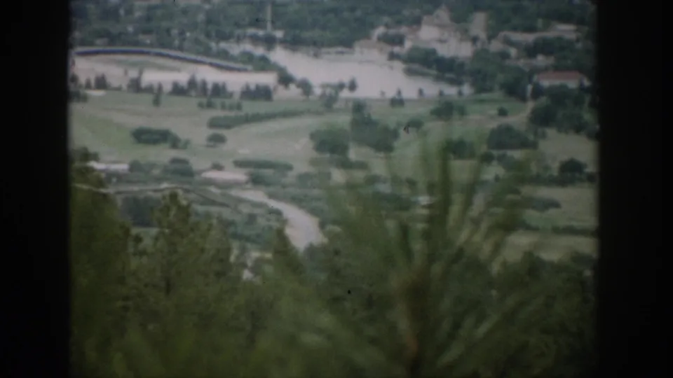 1959 Colorado 8mm frame of a golf course and lake area with roads, buildings, and soft foreground trees behind a dark vertical frame.