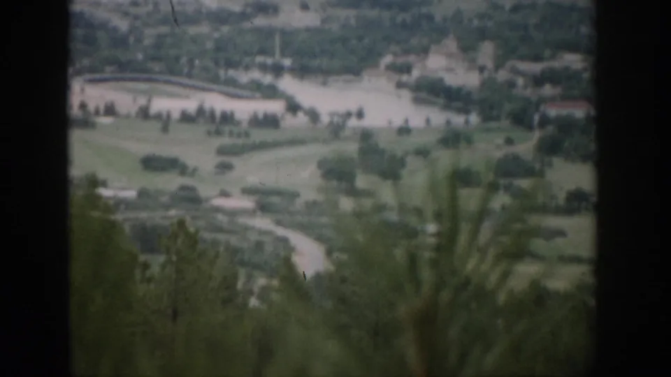 1959 Colorado 8mm frame with a visitor's overlook view across fields, a curved road, water, trees, and distant building shapes.