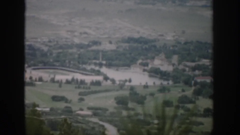 1959 Colorado scenic home-movie frame showing a muted, high-angle valley view with water, open fields, roads, and distant buildings.