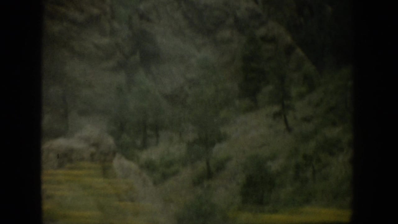 1959 Colorado home-movie frame showing a hazy valley settlement below green slopes, filmed from a moving car.