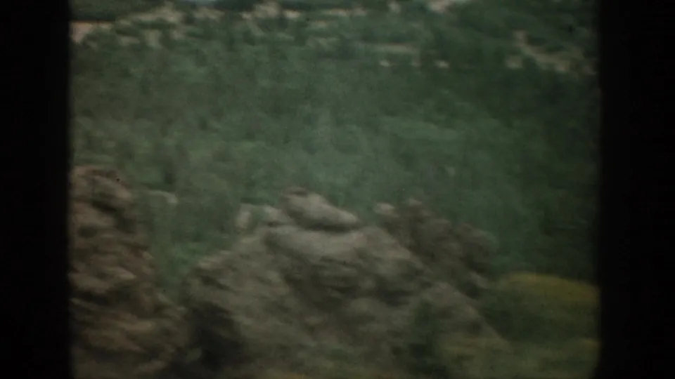 1959 Colorado travel footage frame with rocky outcrops, dense green brush, and soft movement from the car window.