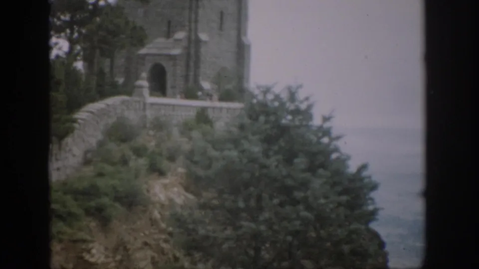 1959 Colorado home-movie frame showing a hillside stone church or tower above trees, with water visible beyond.