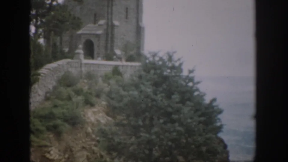 1959 Colorado 8mm frame showing a stone tower, arched doorway, masonry wall, greenery, and hazy horizon.