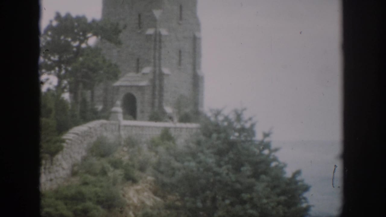 1959 Colorado 8mm view of a multi-story stone tower with arched entry, wall, shrubs, trees, and pale sky.