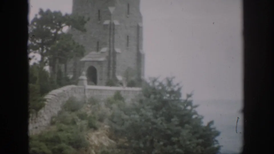 1959 Colorado 8mm view of a multi-story stone tower with arched entry, wall, shrubs, trees, and pale sky.