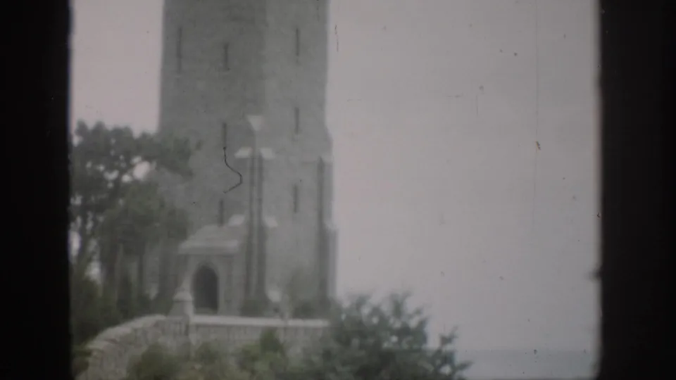 1959 Colorado 8mm frame looking upward toward the stone tower, arched opening, wall, lamp post, and trees.