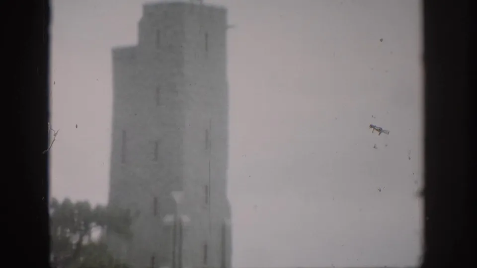 1959 Colorado 8mm frame of the upper stone tower, with a lamp post, treetops, and washed pale sky.
