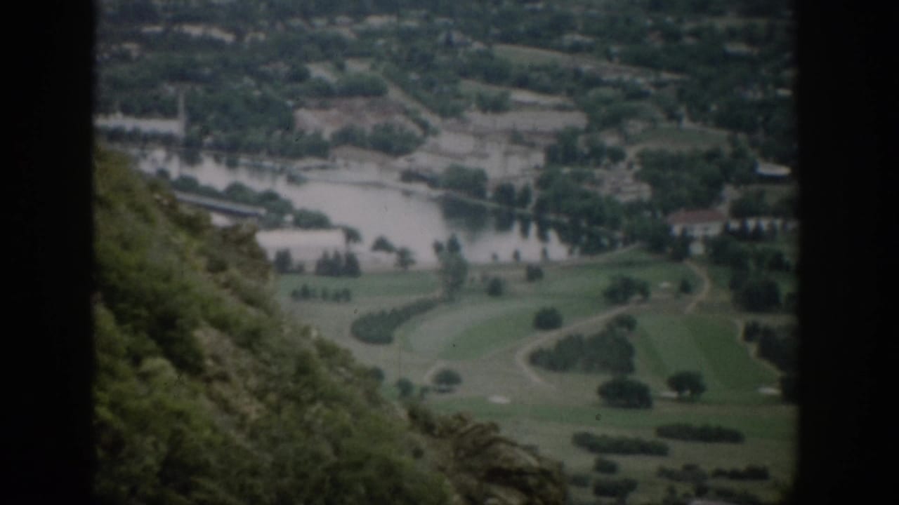 Elevated view over a river, green fields, trees, and distant buildings, framed by dark film borders.