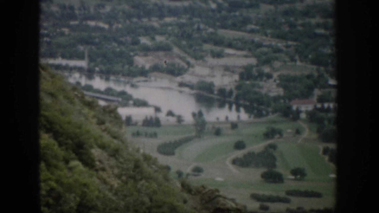 1959 Colorado home-movie frame from an overlook, with a river, open green ground, scattered trees, distant buildings, and hillside vegetation.