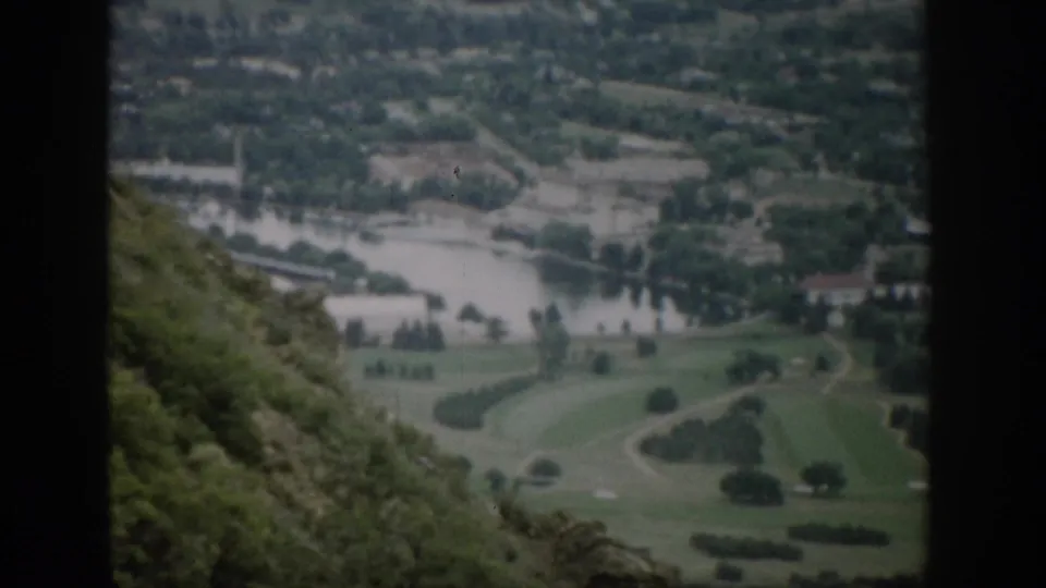 1959 Colorado home-movie frame from an overlook, with a river, open green ground, scattered trees, distant buildings, and hillside vegetation.