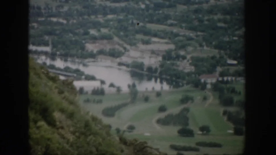 Mid-century Colorado 8mm view looking down toward a river, open fields, small clusters of buildings, trees, and scrubby hillside foreground.