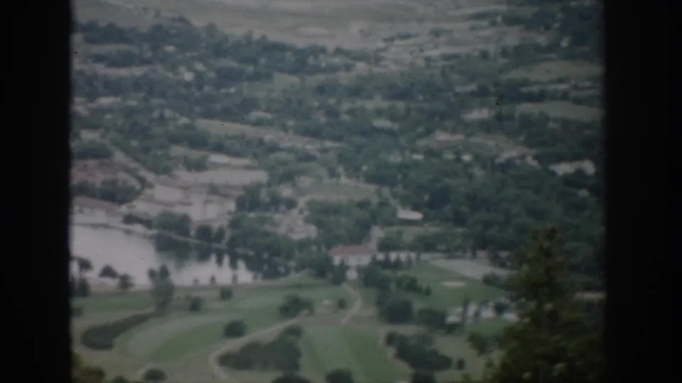 1959 Colorado frame showing a slightly wider overlook of a river valley, open green fields, tree lines, distant town buildings, and vintage film borders.