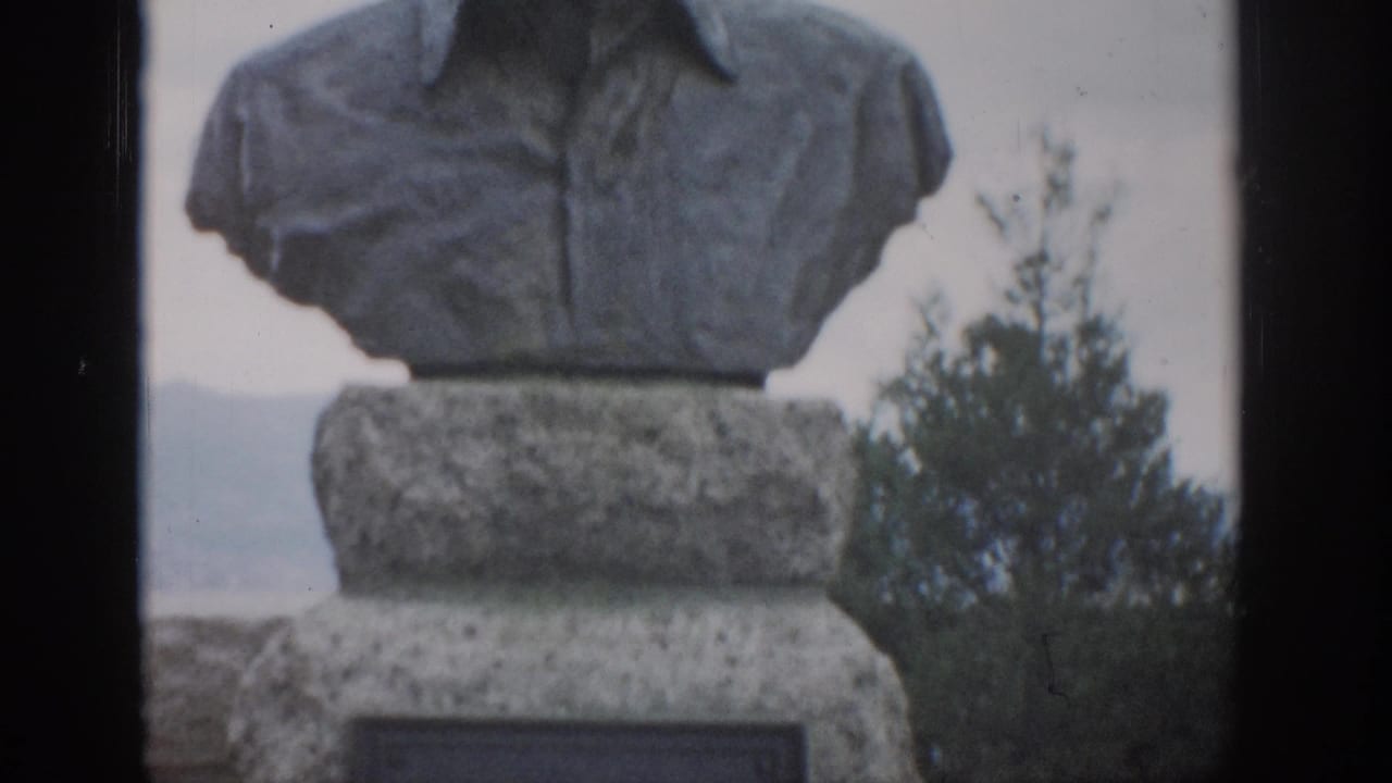 Stone bust monument on a pedestal with plaque, trees, and distant hills behind it.