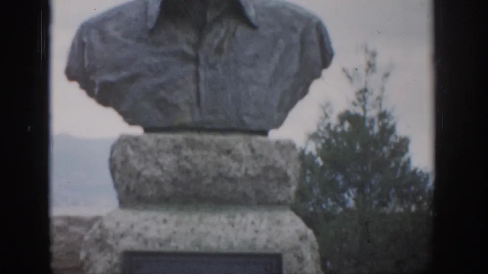 1959 Colorado 8mm frame showing a stone bust monument cropped at the head, with an engraved pedestal, trees, and pale hills in the background.