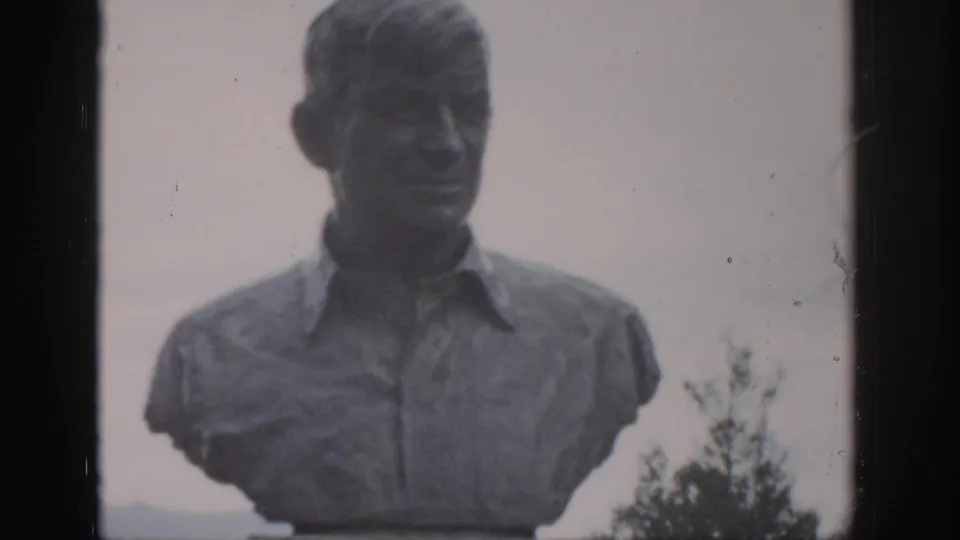 1959 Colorado 8mm frame of a stone bust of a man in a collared shirt, seen outdoors with trees and a hazy hillside behind.