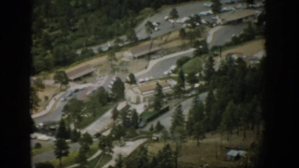 1959 Colorado 8mm frame looking down over a park-like area with roads, small buildings, parked cars, trees, and dense forest nearby.