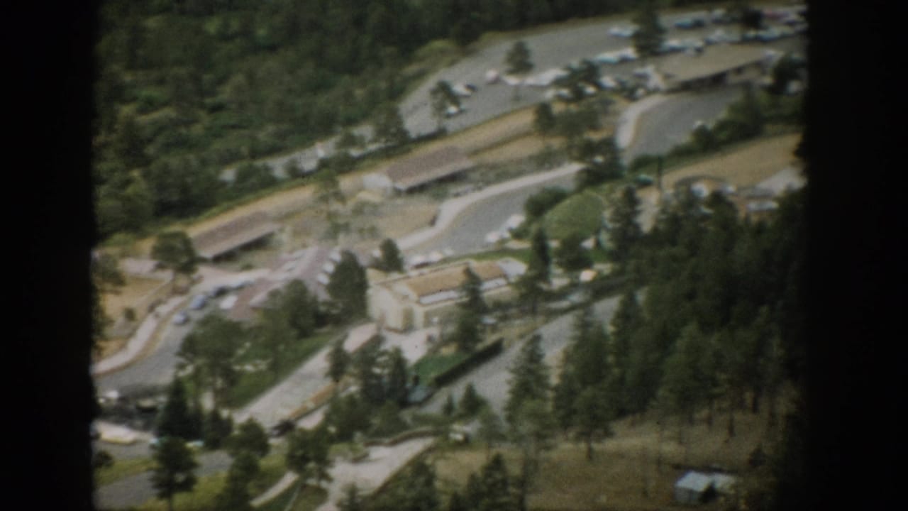 1959 Colorado 8mm aerial view of a small town with winding roads, parked cars, low buildings, and dense pine forest below.