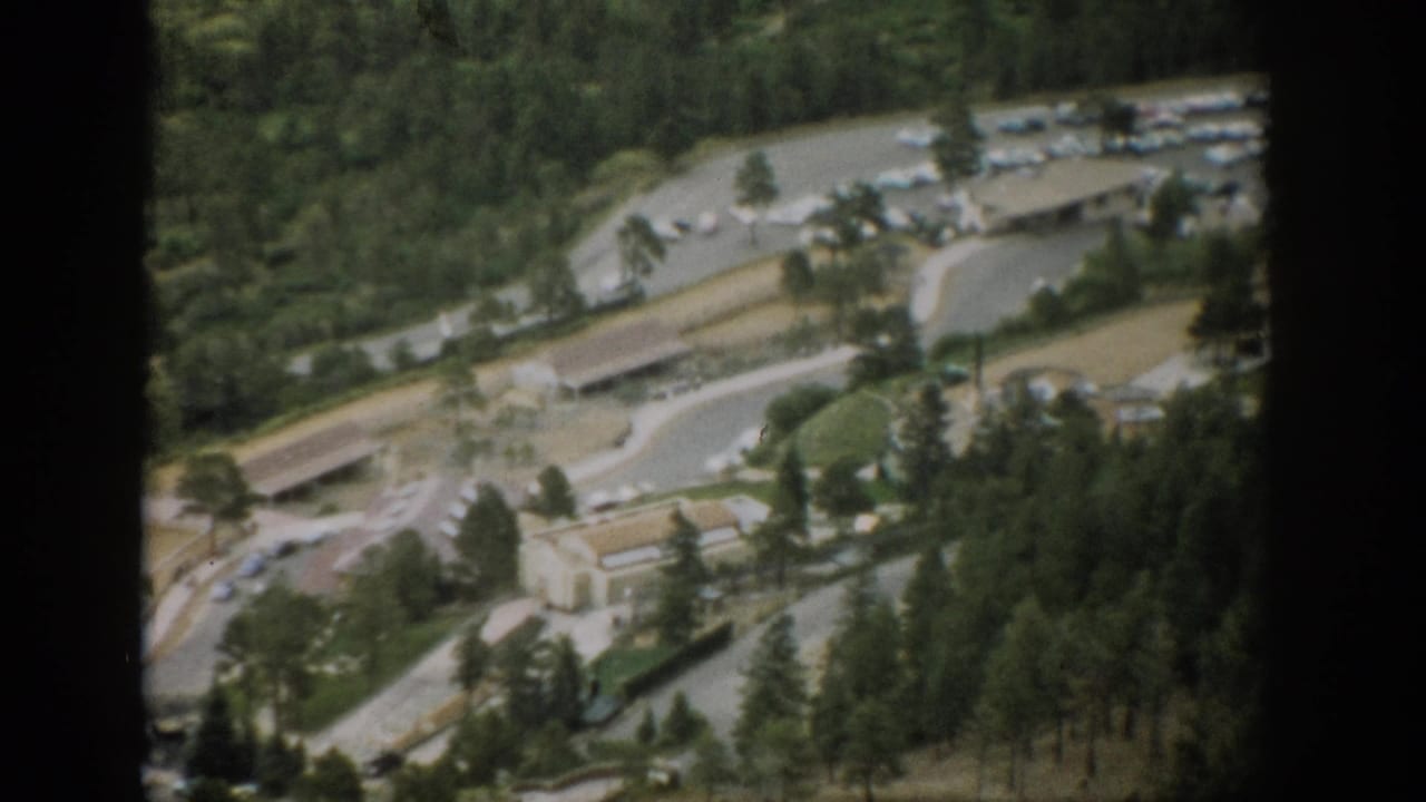1959 Colorado aerial home-movie frame showing light traffic infrastructure, parked cars, flat-roof buildings, and pine-covered terrain.