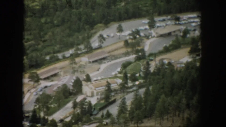 1959 Colorado aerial home-movie frame showing light traffic infrastructure, parked cars, flat-roof buildings, and pine-covered terrain.